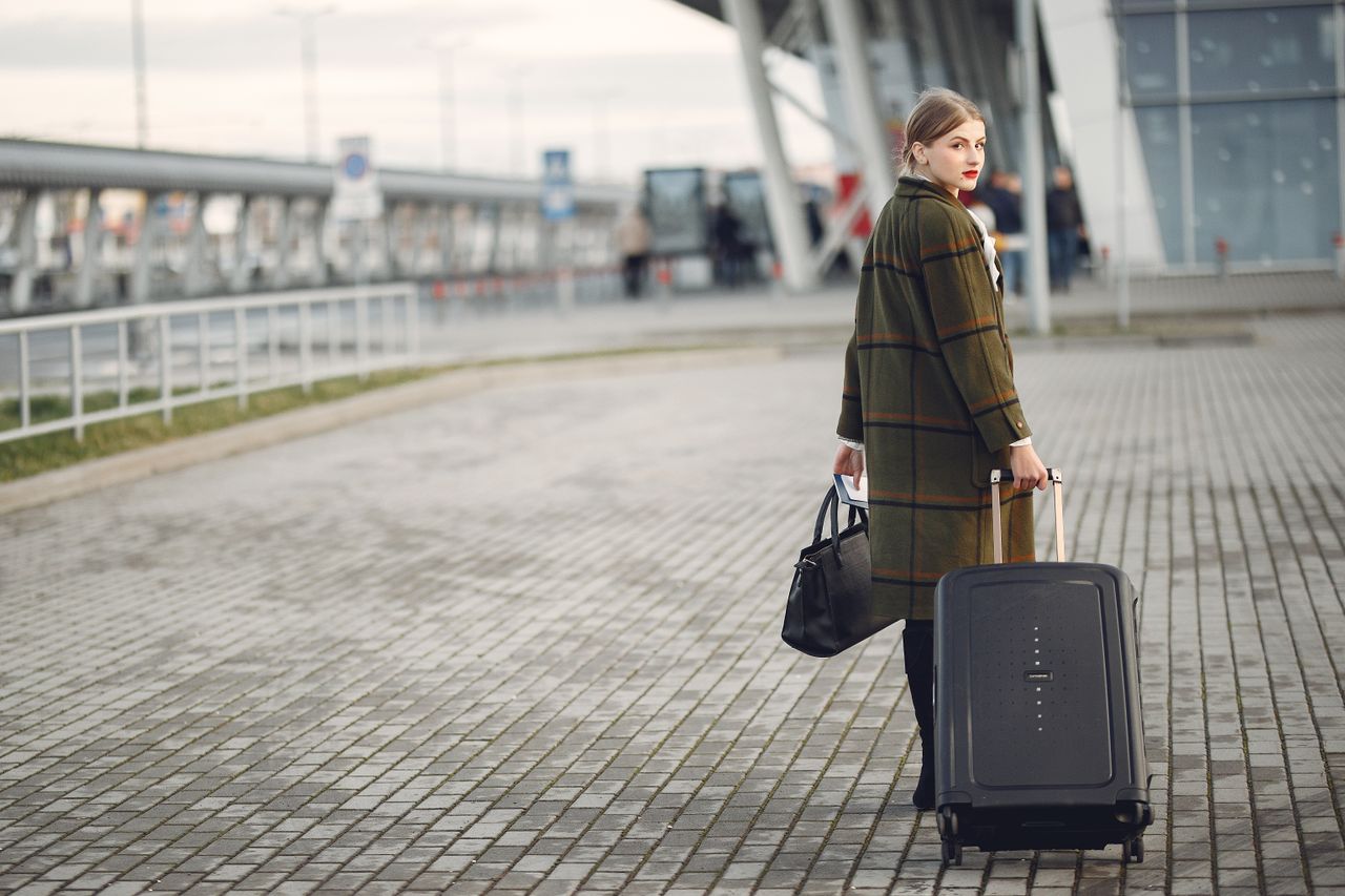 services-01 Full length of stylish female traveler in warm coat walking with suitcase and bag looking at camera after arrival against airport terminal
