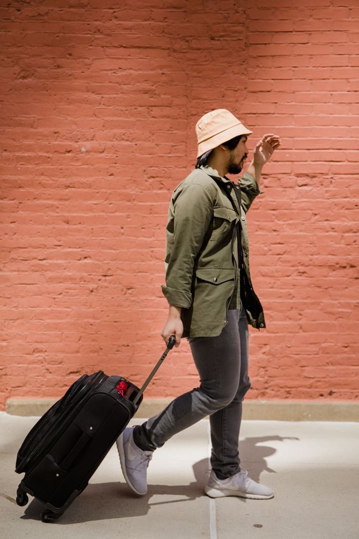 services-02 A man wearing a green jacket and hat pulls luggage past a brick wall.